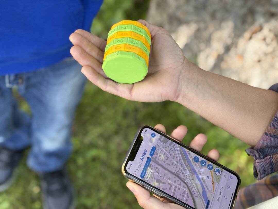 Ein vierstelliges gelbgrünes Zahlenschloss liegt auf einer ausgestreckten Hand, daneben sieht man ein Mobiltelefon mit Geodaten. Im Hintergrund ist verschwommen ein Baum und ein Mensch zu sehen.